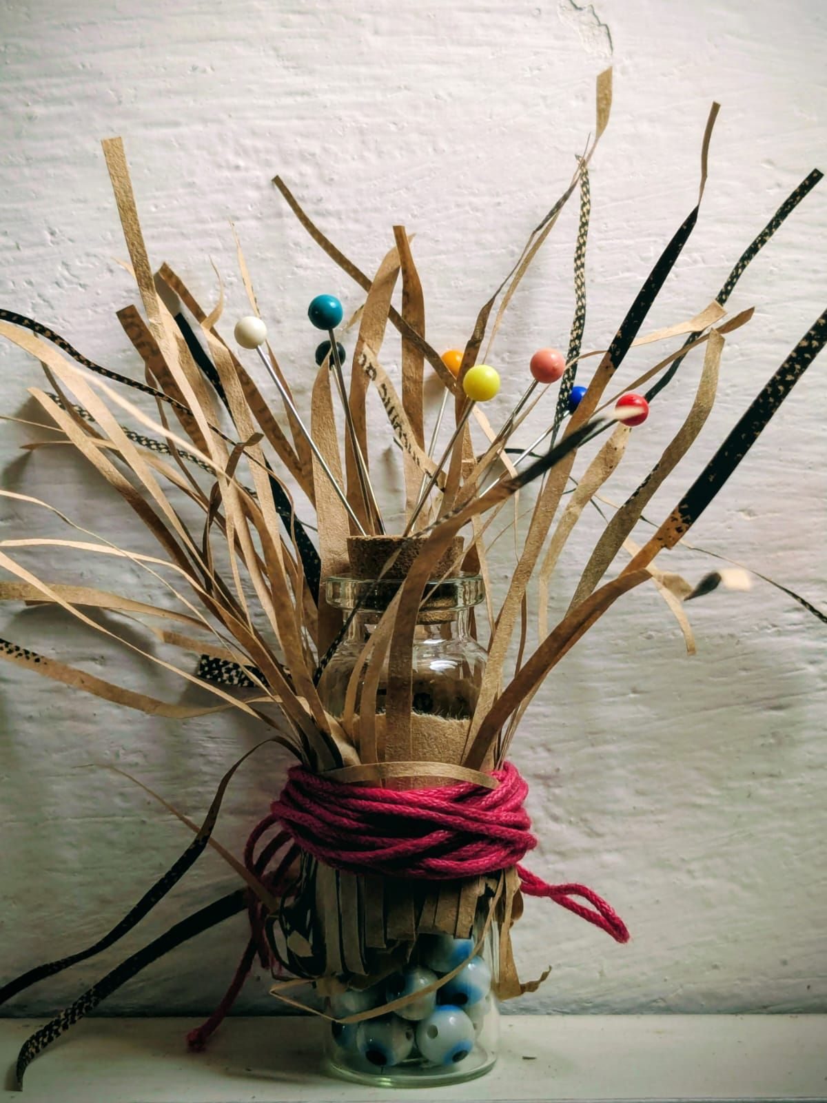 A glass bottle containing glass bead eyes, with sewing pins planted in the cork top, wrapped with shredded brown paper with red yarn tied around to keep it in place