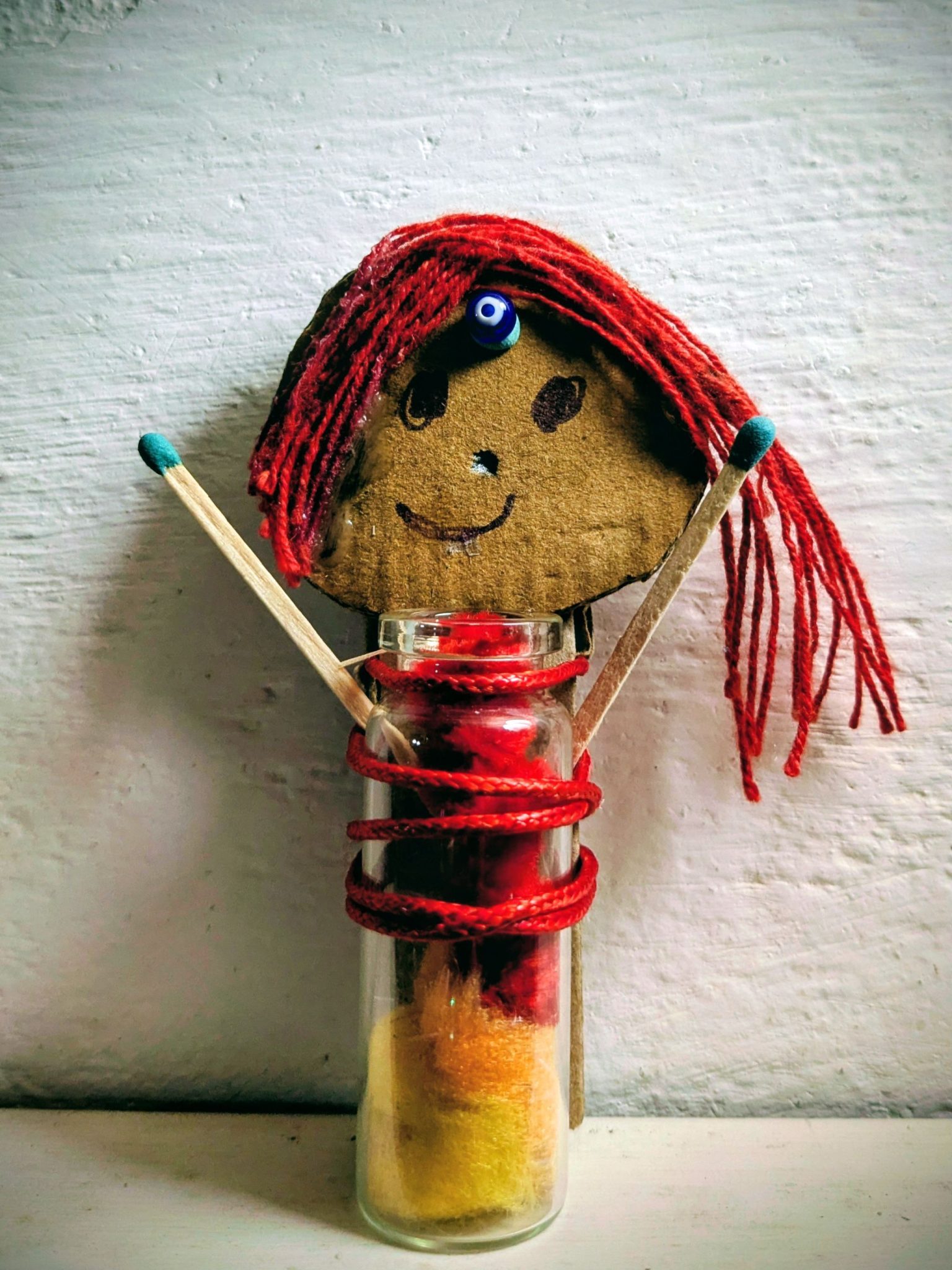 A small glass bottle with colourful pompoms in it, a face drawn on cardboard with red thread hair, a blue bear as bindi, 2 arms made of matchsticks