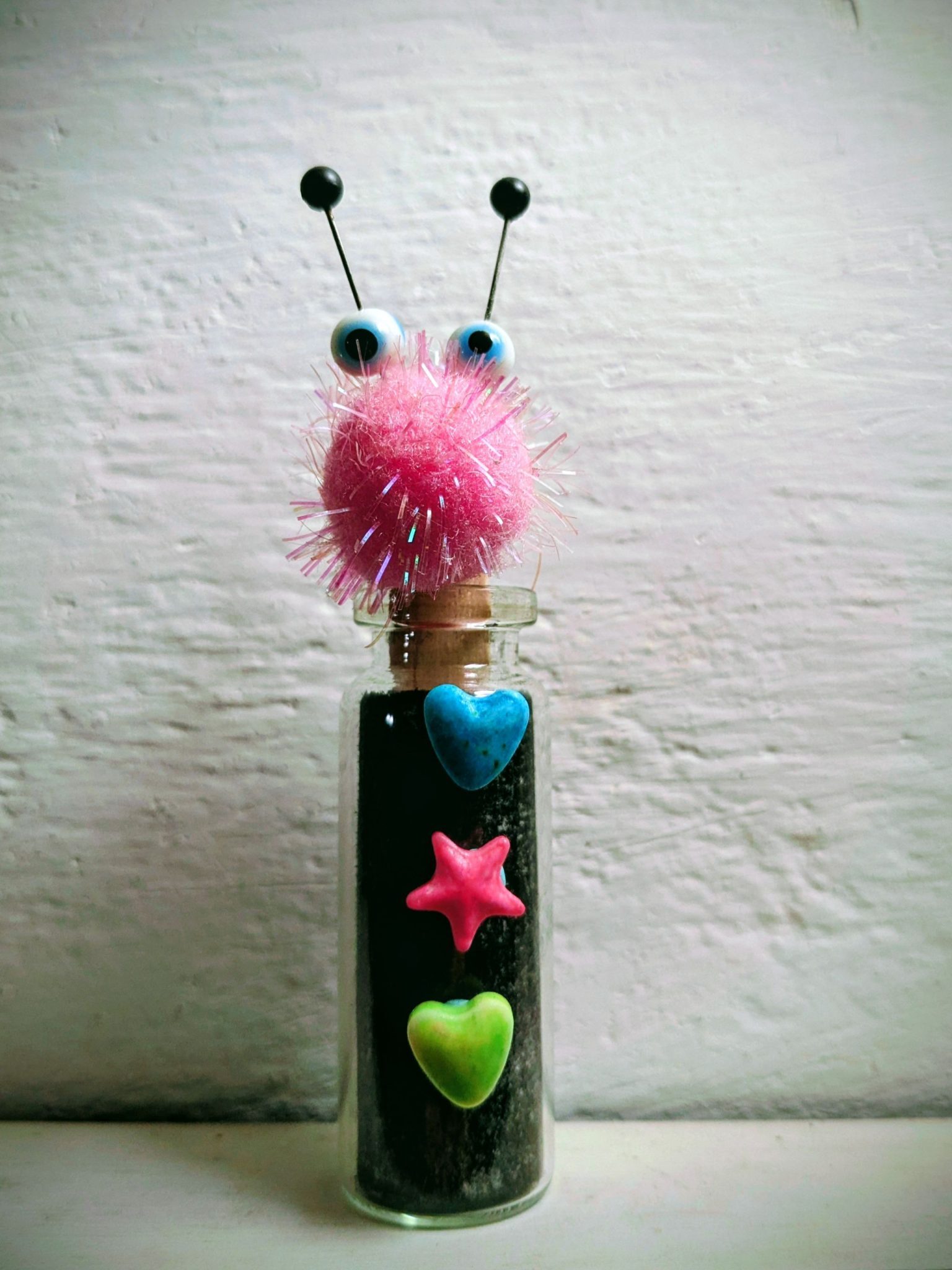 A small glass bottle containing a black charcoal drawing with 3 candies as button and a head made of a sparkling pink pompom, and 2 glass eyes held by pins that look like antenna