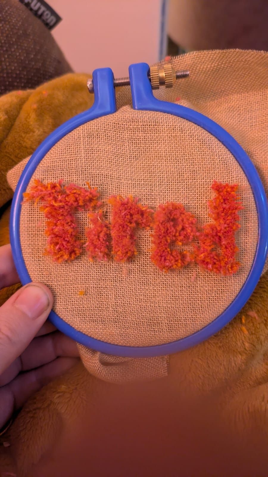 A brown linen in a blue embroidery hook inside the hoop the word tired is spelt out in 3d pink and orange punch needle