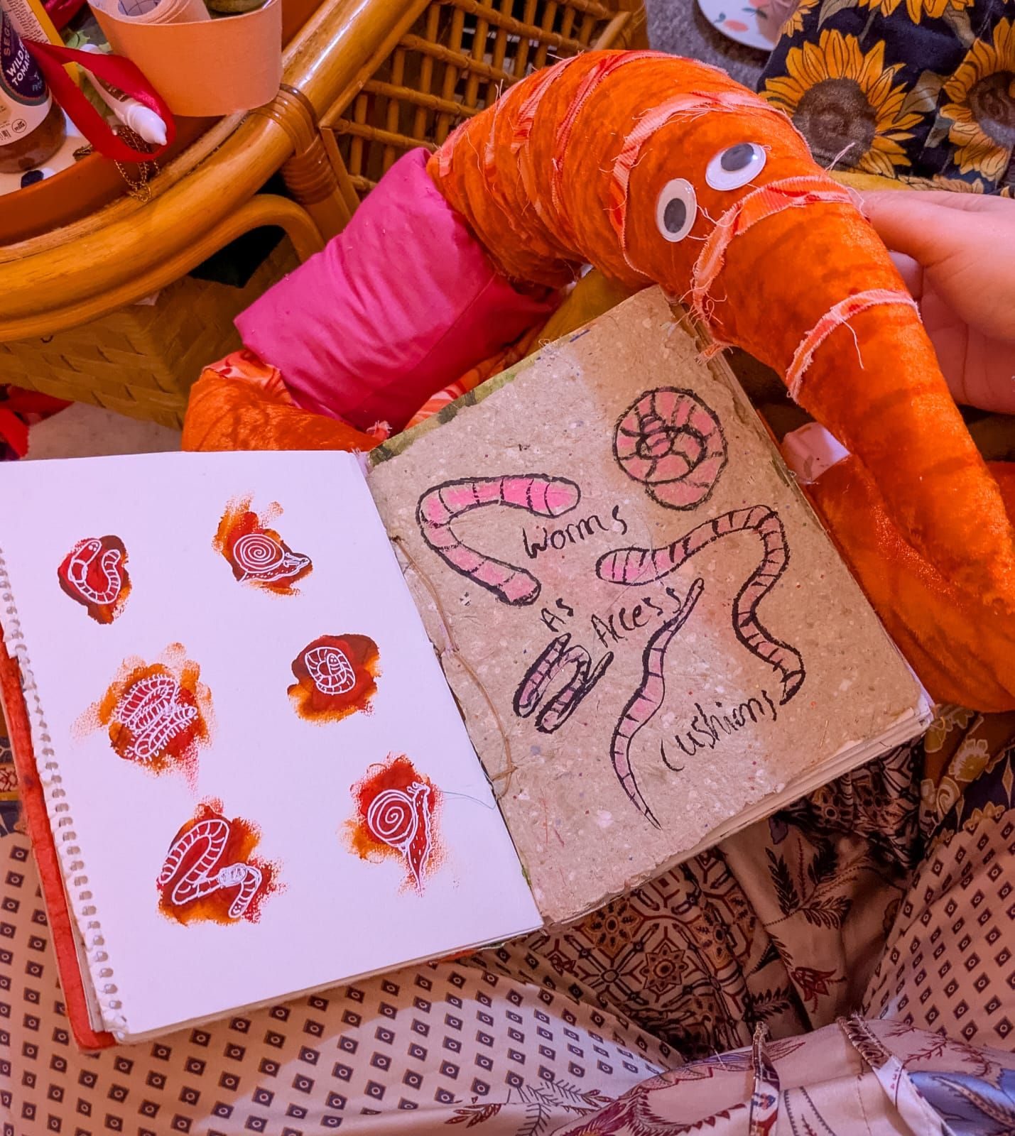 The image is of a sketchbook with drawings of worms, snails and millipedes on one page and another page with drawings of worms as accessible cushions. In the background of the photo there is also a fabric worm.