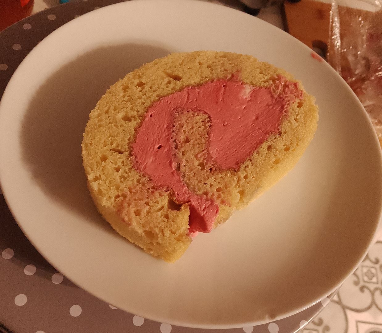 A photo of a slice of Swiss roll on a white plate. The plain sponge is rolled up with a pink cream filling.