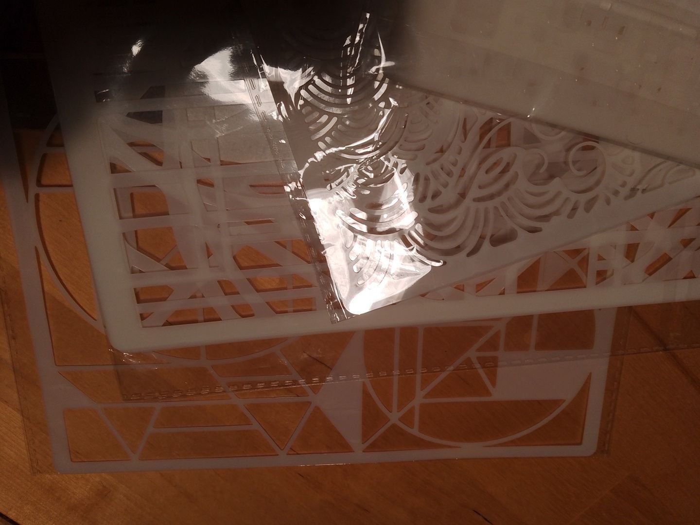 A photo of several white plastic stencils overlaying each other on a wooden tabletop. The stencils are in shiny cellophane wrappers and the overhead light is reflecting off a patch of them, making them look silver.