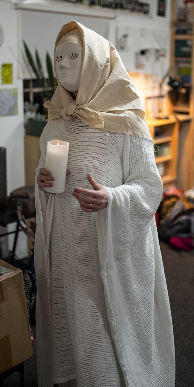 A woman wearing a white mask covered with paper lace. She is wearing a cream cotton headscarf, and a long cream dress under a white robe made from a hospital blanket. She is carrying a lighted pillar candle.
