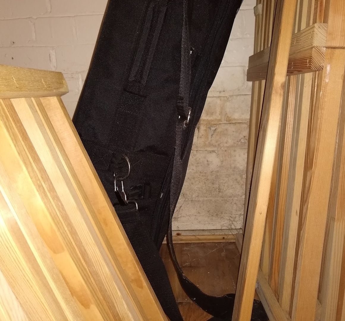 Some dismantled wooden shelving units and a black nylon gig bag in a white painted brick cupboard space. There are cobwebs and damp patches on the walls.