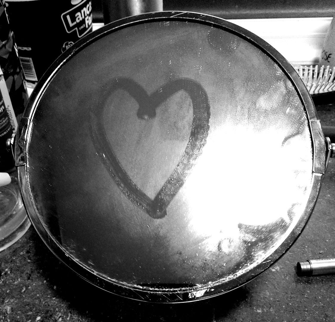 A black and white photo of a round silver-framed bathroom mirror. The mirror is steamed up and there is a heart shape drawn in the condensation with a fingertip.