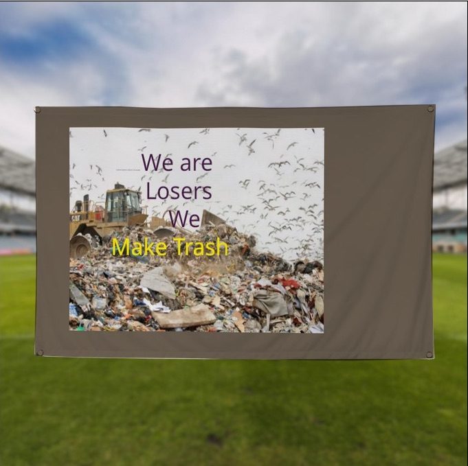 a pile of landfill being moved by a digger, with scripting purple saying 'We are Losers  We' in purple, below it reads 'Make Trash' in yellow, the flag has a background of muddy brown, it appears to be held up infant of cloudy blue sky, there is grass below and there and there are signs of a stadium of some sort suggesting this is a team, one with supporters