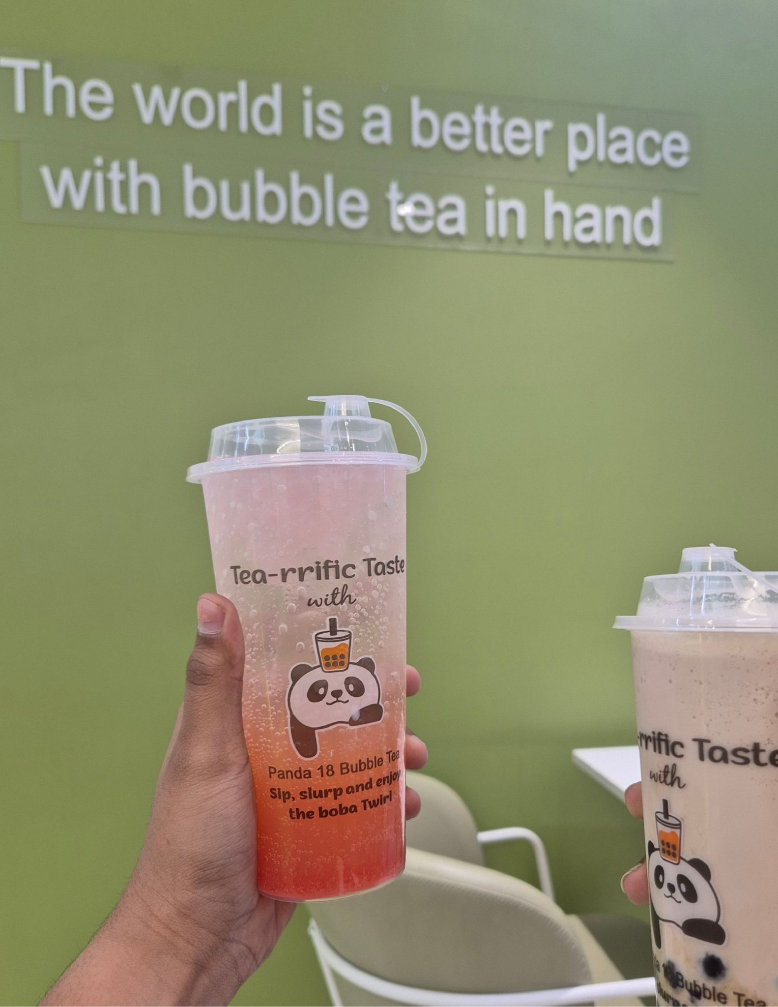 Two hands raise their bubble teas beneath a hopeful green wall, capturing a simple, sweet moment shared with someone you love.