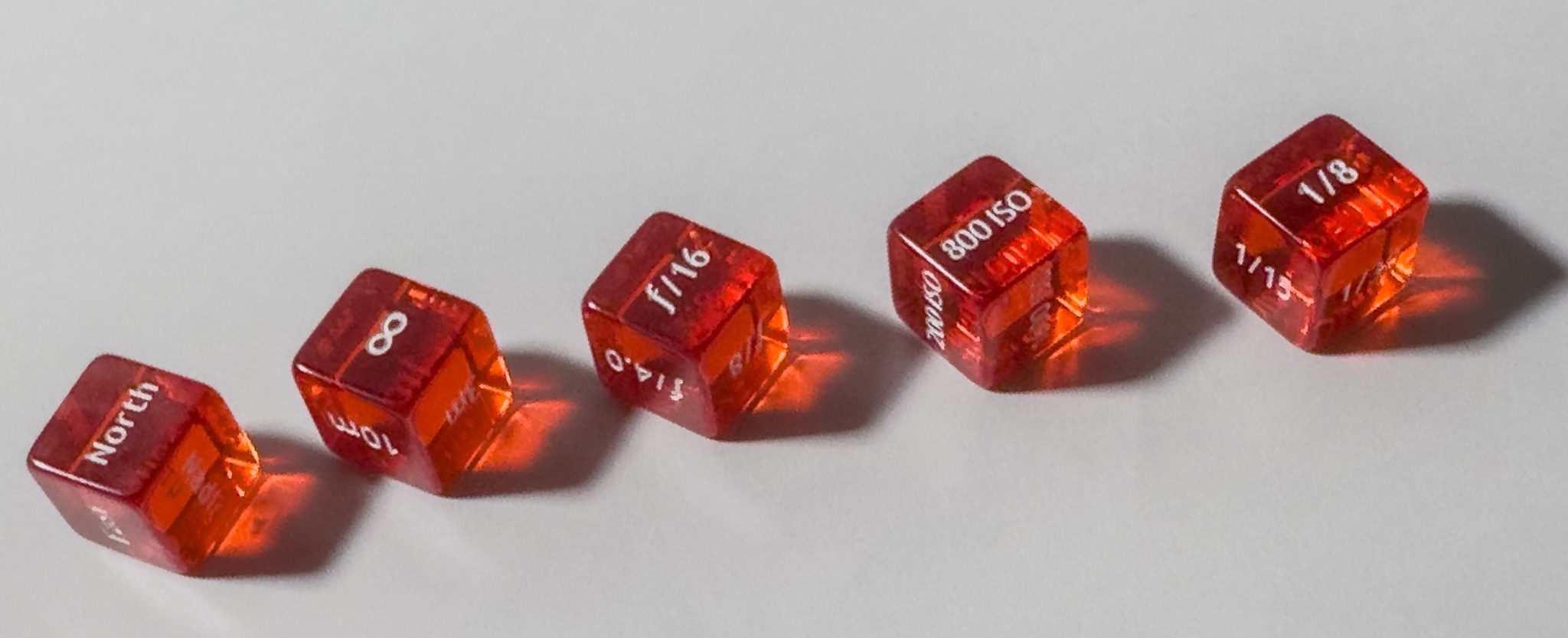 5 red dice, each one with a different photography variable, shutter speed, aperture, ISO, focus distance and direction.