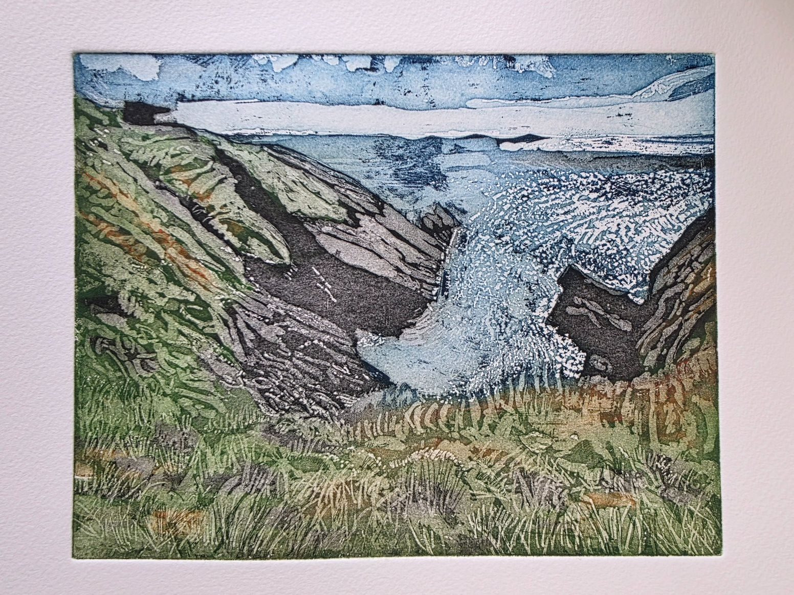 Coastline with black rocks going into the sea. Grass in the foreground. Blue sky and sea with sun glittering on the water