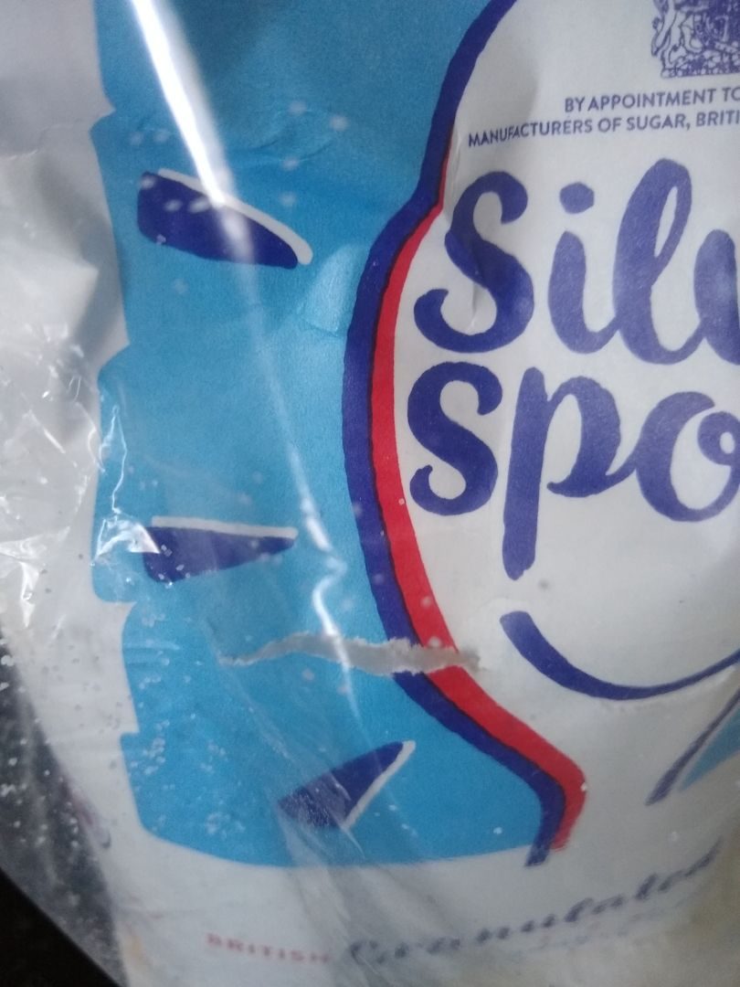 A close-up photograph of a white and blue Silver Spoon paper sugar bag inside a plastic bag. The paper bag has a tear in it, and there are stray grains of sugar clinging to the inside of the plastic bag.