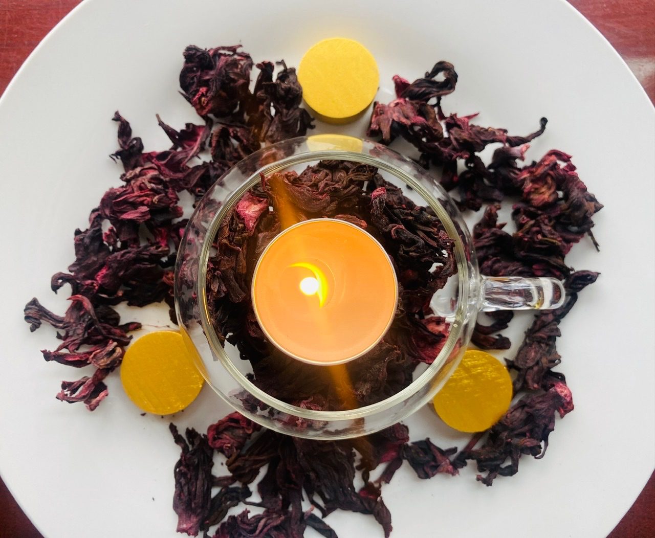 Photograph of a white plate with a clear cup filled with dried hibiscus flowers inside and overspilling onto the plate. There is a lit incense stick in the cup, between the flowers.