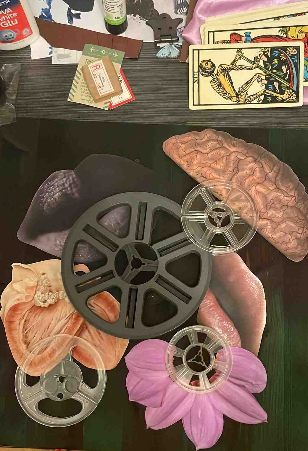 An overhead shot of a cluttered craft table shows tarot cards, glue, scraps of paper, and film reels arranged over a collage of flowers and anatomical brain imagery.
