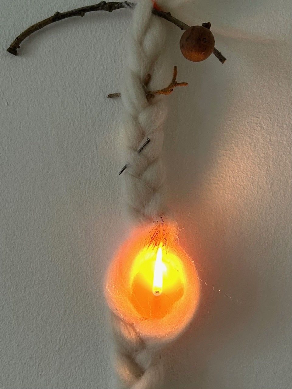 Sheeps wool plat hanging on a wall with various bits woven into it- an oak gall on a twig, a nail, and at the bottom a lighted bees wax candle