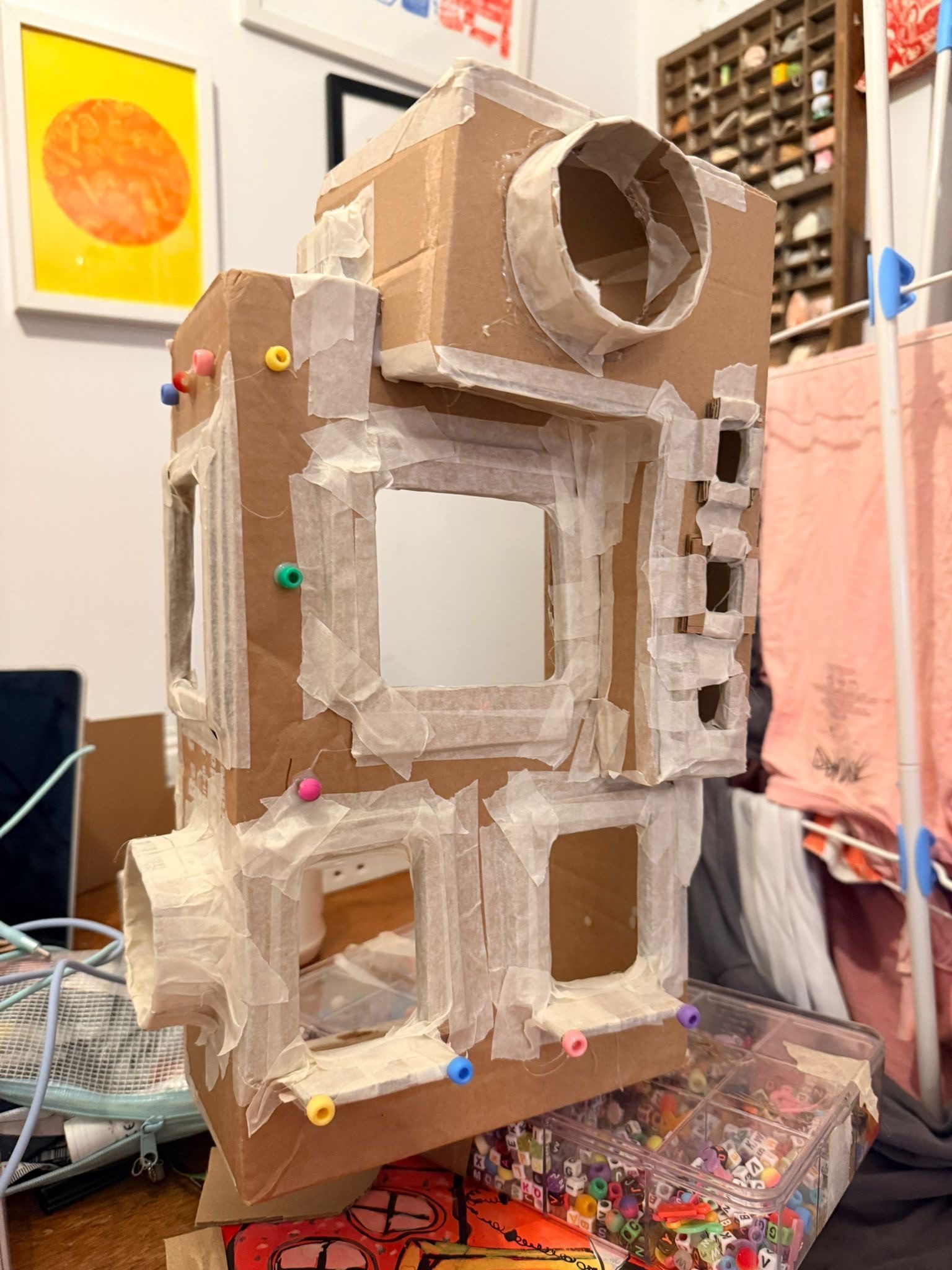 a photo of a cardboard sculpture resembling an abstract building with various windows. its held together with making tape, and also has some colourful beads stuck to it!