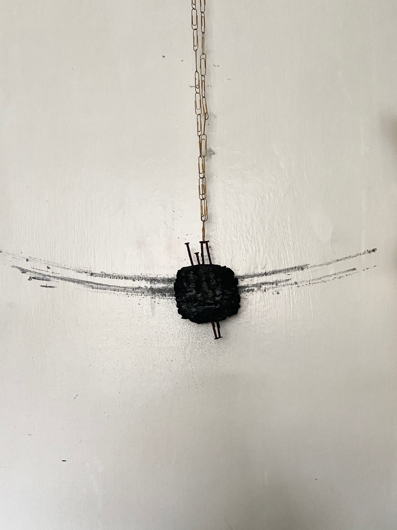 a charcoaled piece of wood with nails, swinging on a necklace of gold paperclips, drawing marks