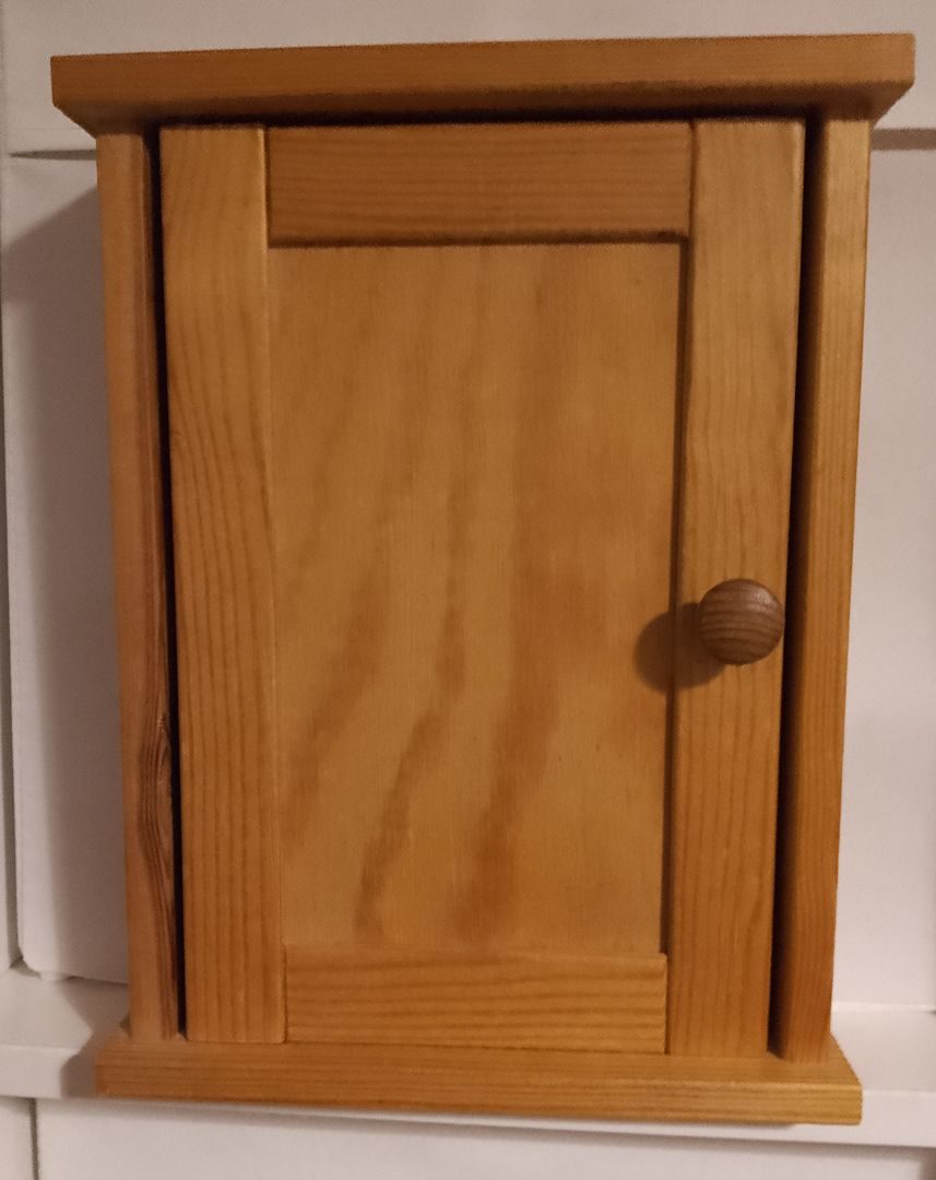 A small wooden key cabinet with its door closed so you cannot see what is inside. It is made of pine or a similar light wood, and is about 27cm high, 20cm wide and 8cm deep. The small door knob is on the right-hand side of the door.