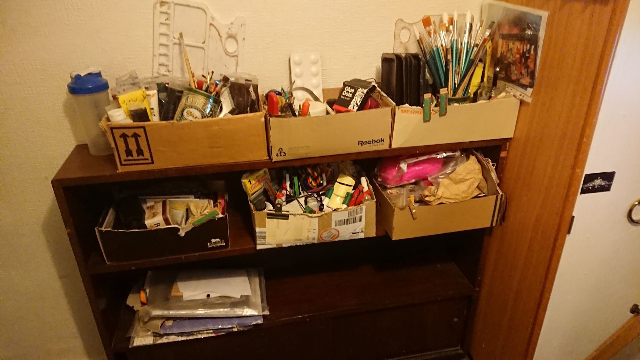 A photo of shoe boexs containing art equipment on a book shelf , so things like paint brushes , paints pens etc