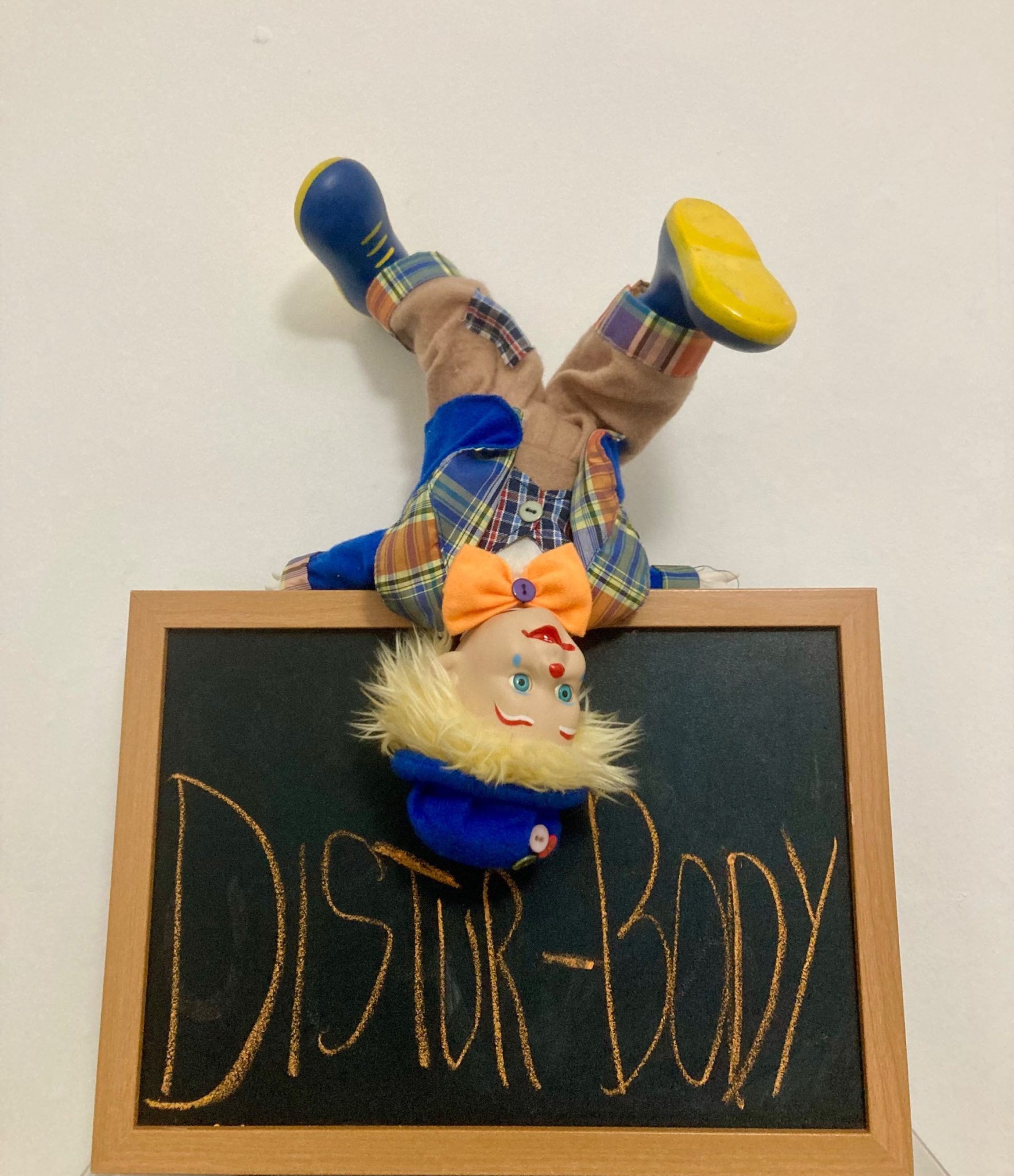 A photo of a clown in an upside down position, balancing with their shoulders above a blackboard that shows the text 'Distur-Body' written on it