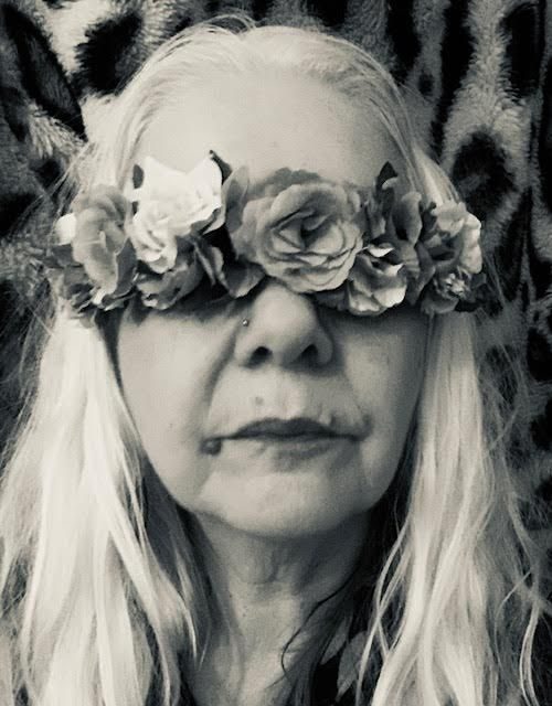 [black and white photo of a face with long white hair and a blindfold of flowers, against a leopard print fur background]