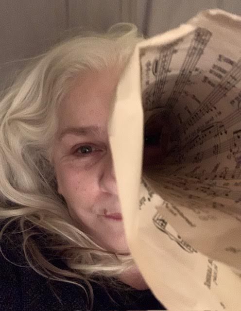 [colour photo of a face, long white hair, a piece of sheet music rolled into a cone and held up to one eye, the sheet music obscuring the eye and most of the right side of the face