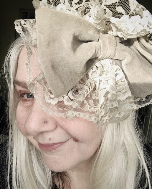 colour photo of face in half profile, smiling, one eye visible, lace rosette on grey half-hat with grey velvet bow over the other eye