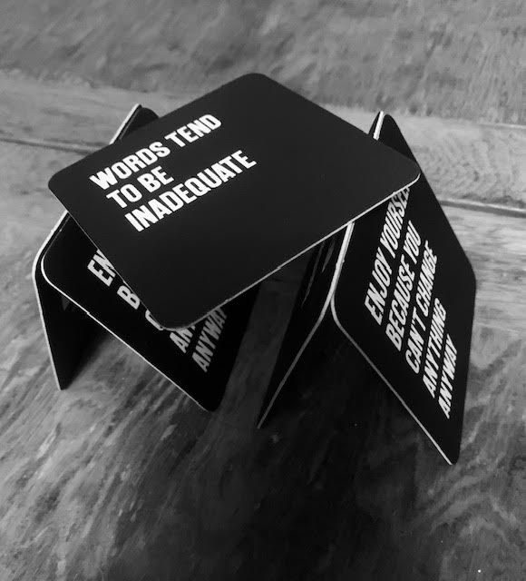 black and white photo of five drinks mats with text on them, two pairs of two forming triangles and the fifth balanced at an angle on top of the apexes of the triangles. The top card reads: Words tend to be inadequate’