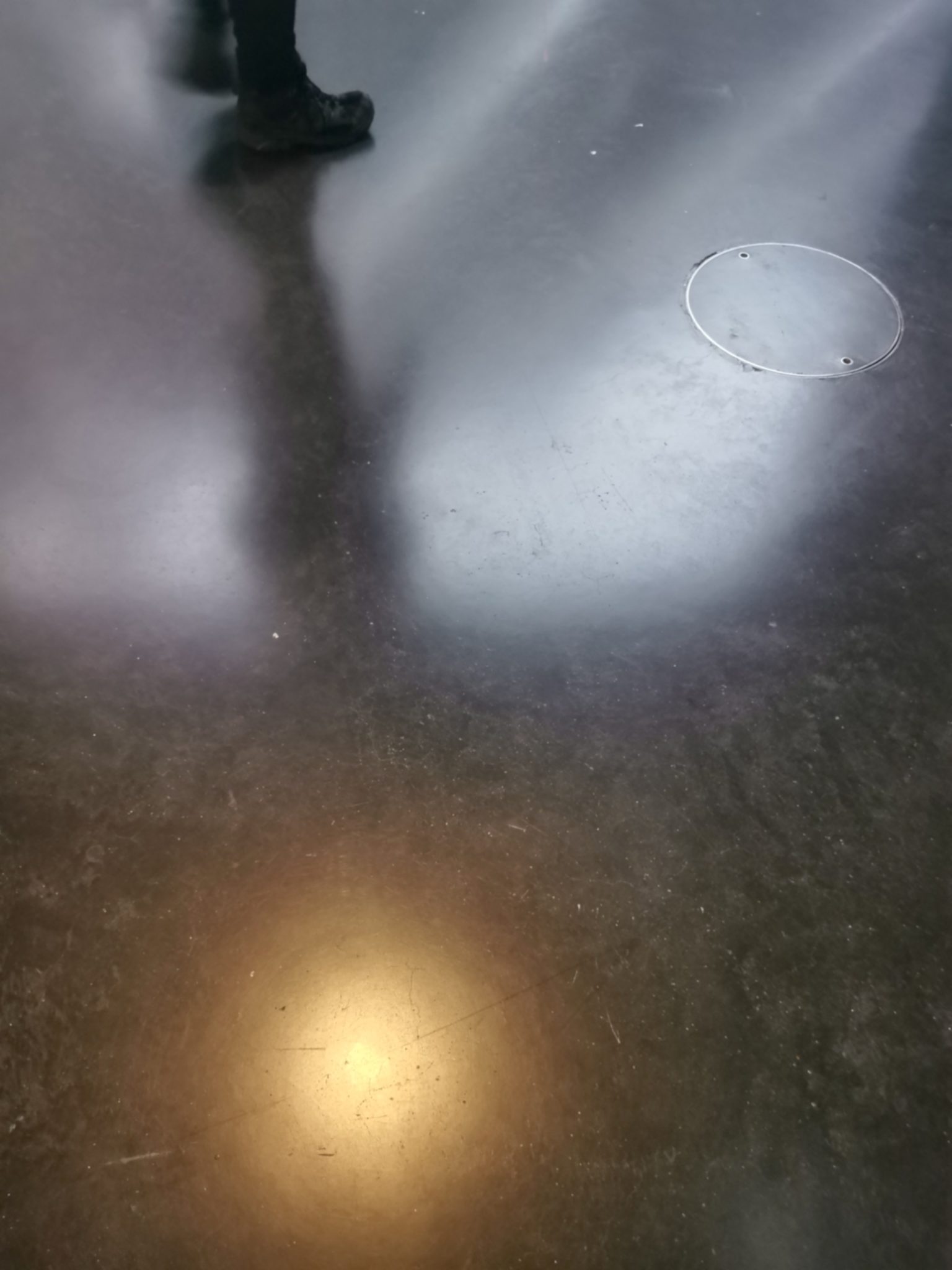 Photo taken in a gallery with a light from a screen reflecting off the highly polished floor and a persons feet and legs in black trousers and shoes reflected as they walk past at top of photo.