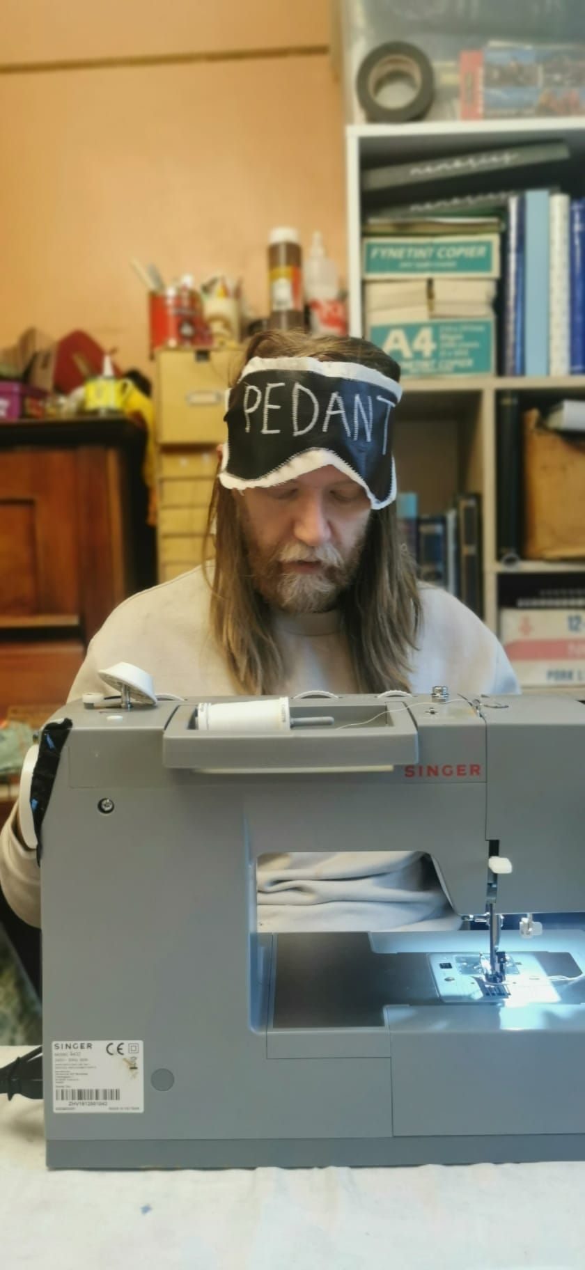 A photo of the artist, working at a sewing machine wearing a blindfold not covering their eyes.  The word 'pedant' is written on the blindfold