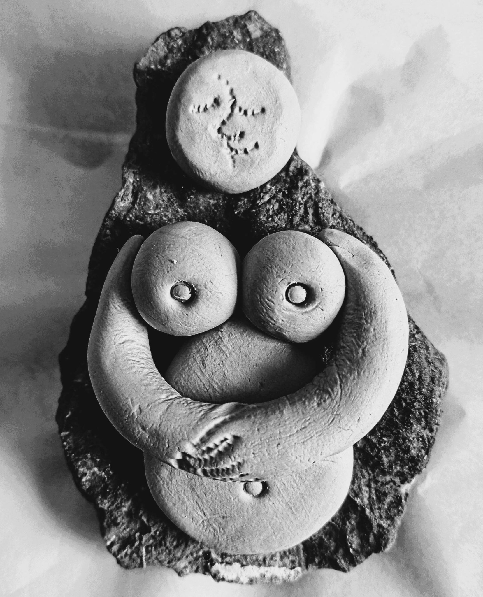 Rectangular photographic image of an exaggerated female torso comprised of smooth clay anatomical features on a rough stone base, with a full moon face