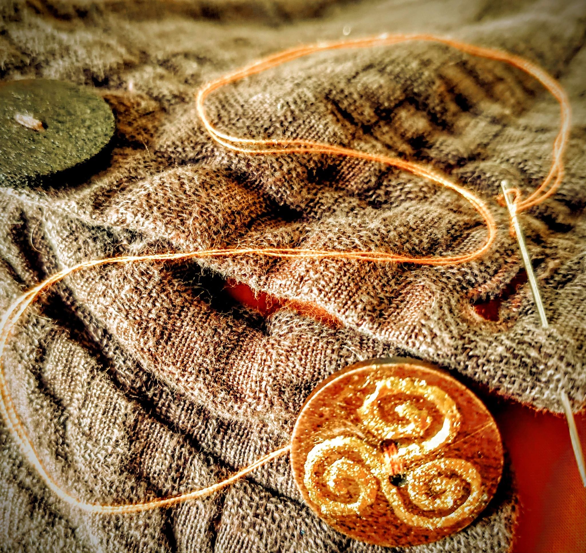 Square, photographic image of a section of textured fabric and 2 wooden buttons, one of which has just been sewn back on with contrasting bright thread and has been embellished in gold with a Celtic triple spiral symbol. The needle is still attached to the thread.