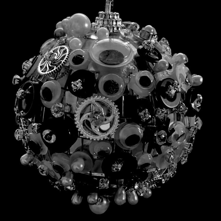 A black and white image of a disco ball which is covered in buttons, cogs, rhinestones, pearls, and googly eyes