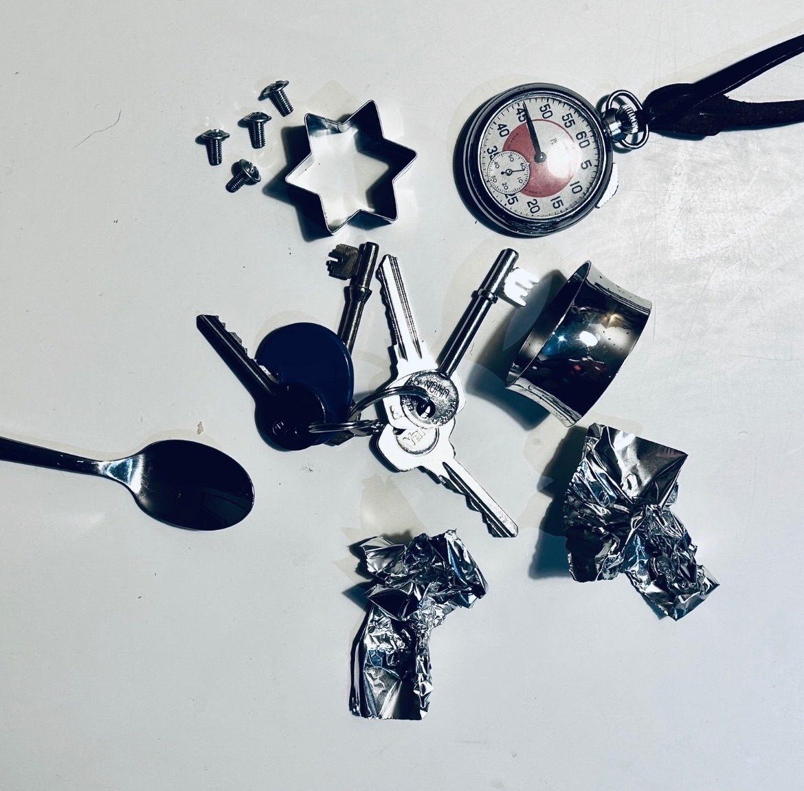 Keys, a spoon, silver foil, screws, pastry cutter, napkin and watch laid out on a white table top