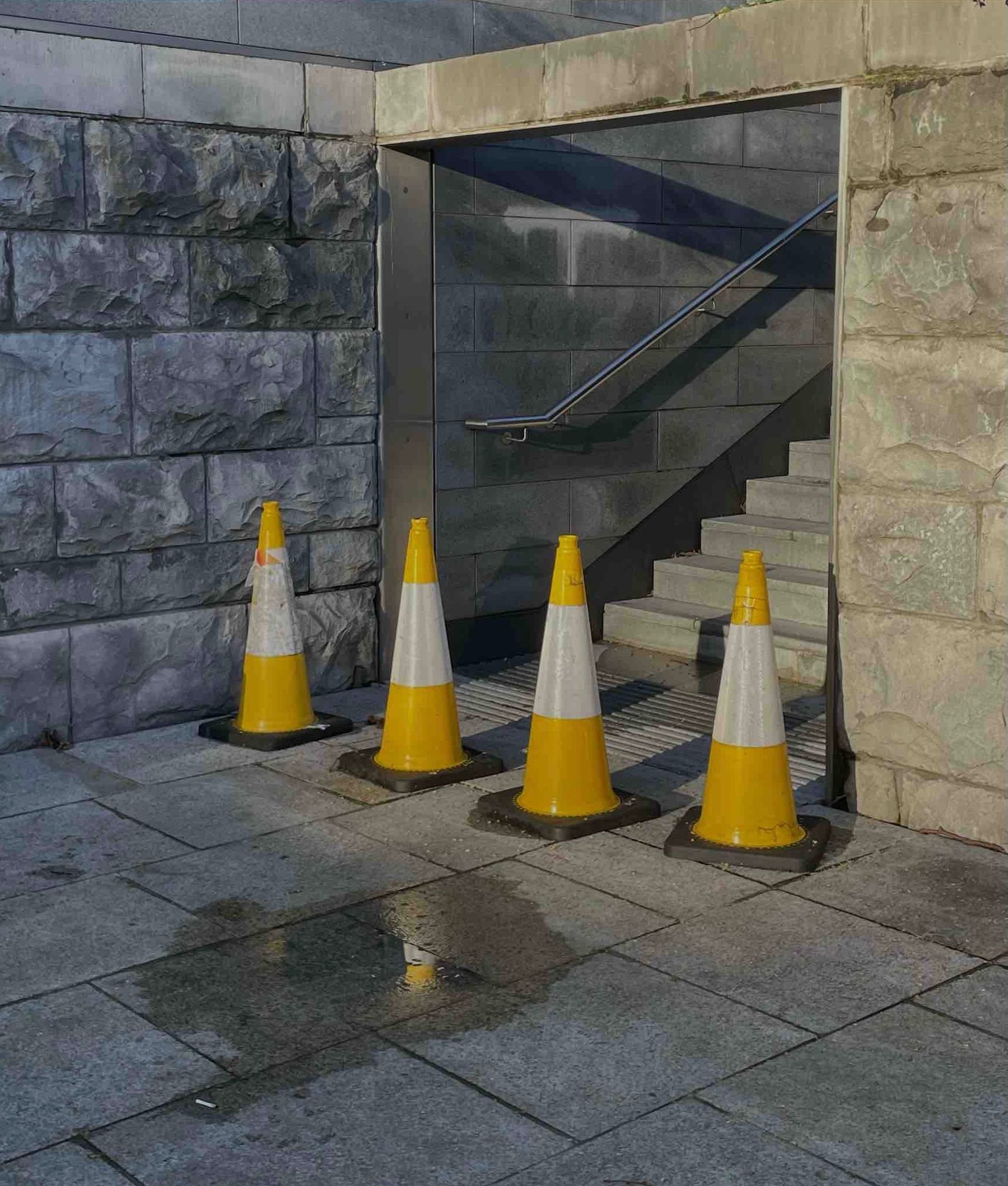 Photo of 4 yellow traffic cones at the foot of a stairwell with grey brick and tiles behind