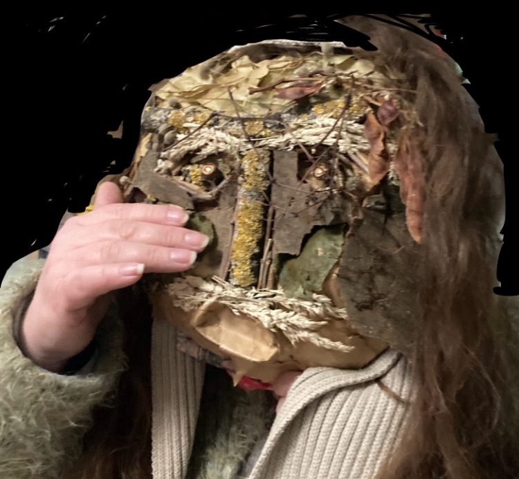 There is a mask in front of my face the mask is of twigs, leaves, seed pods there is tree bark building up features of the cheek bones and contours, there is grasses with seeds that are about the mouth as a moustache and used also for eyebrows, there is branches the have been cut cross sectioned as pupils of the eyes, the nose is a branch with green and blue lichen, the hair is of a few strands of hay, and there is also leaves that help to create more character to the mask. In this photo I am holding this mask so you can see a human hand looking like it is wiping a tear away, there is a beige colour scarf and long hair hanging down beside the mask and it is green colour generally with muted brown tones from the bark and seeds and all natural materials used to make this “green person mask” it is titled “A Little Green Around the Gills, Feeling Sad For Mother Earth today”