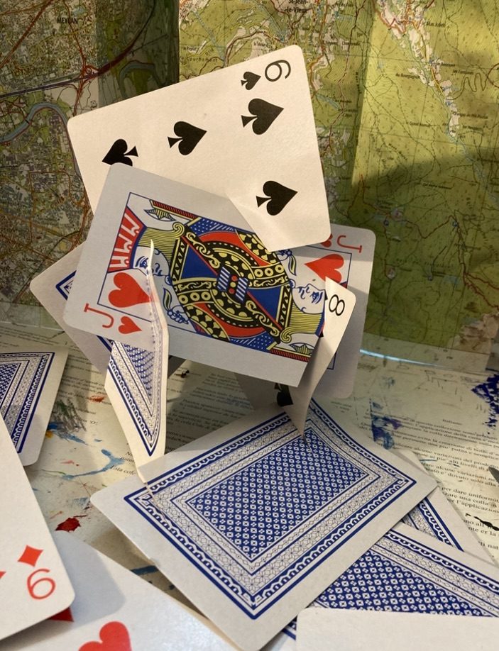 It is 5 playing cards that have been cut into in two places and are in precarious positions slotted into each other and at angles that resemble a random shape. There is the 6 of clubs at the top bridging a gap between two cards that is also sitting cut into a Jack of hearts at the corner a black 8 is showing and these are all slotted into with cuts two more sitting at a right angle this is all balancing. The background is a map that isn’t important but is a back drop to the picture. The title is House of Cards.