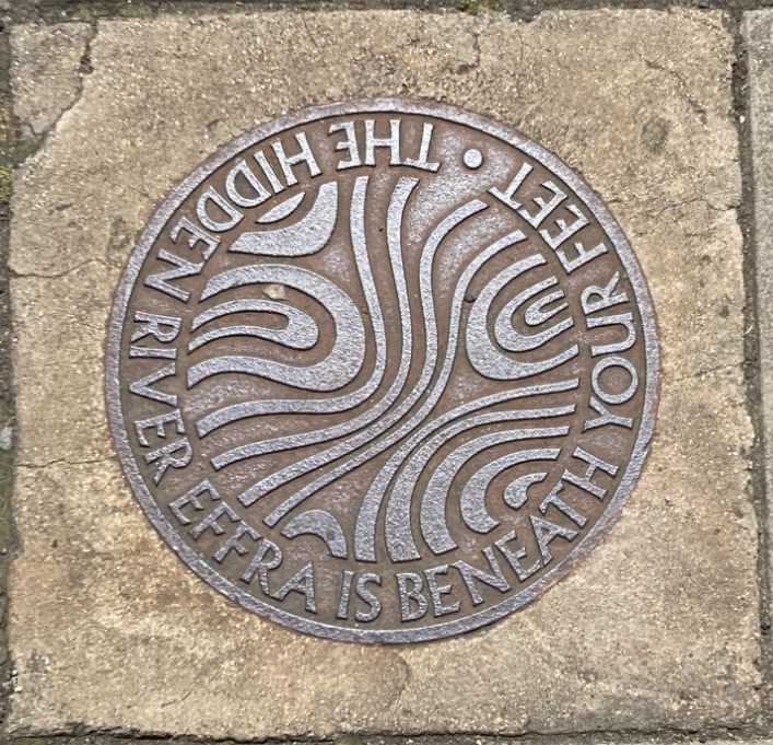This is a picture of a water cover in the street it says The Hidden River Effra Is Beneath Your Feet around the edge of the metal road cover it is circular and in the space between is swirled suggesting moving and swirling water