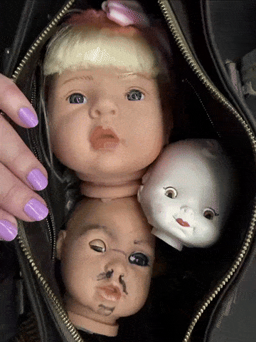 An animated gif showing an open bag with fingers with lilac nail varnish holding the bag open and inside the bag dolls faces are constantly changing, baby dolls heads, a mannequin head, a sex robot head and a woman’s head