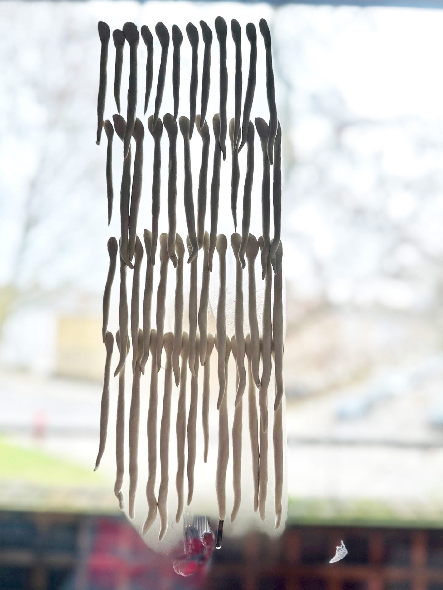 I formed coils of clay by hand and pressed their upper edges onto the window, letting the glass hold them in place. Almost immediately, the clay began to breathe its moisture onto the surface, a faint bloom spreading across the glass. Within moments, droplets gathered and traced slow paths downward. The clay softened in response, loosening its grip, hovering on the verge of slipping free from the window.