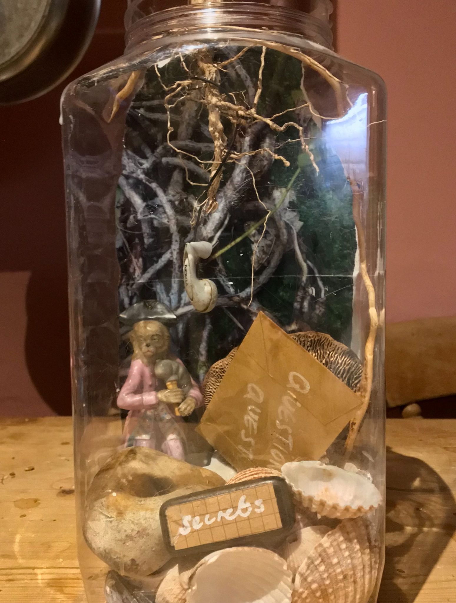 large plastic jar filled with from the bottom -shells, a figurine of a monkey, picture of roots, a small tin with the word secrets and an envelope with the word question and quest
