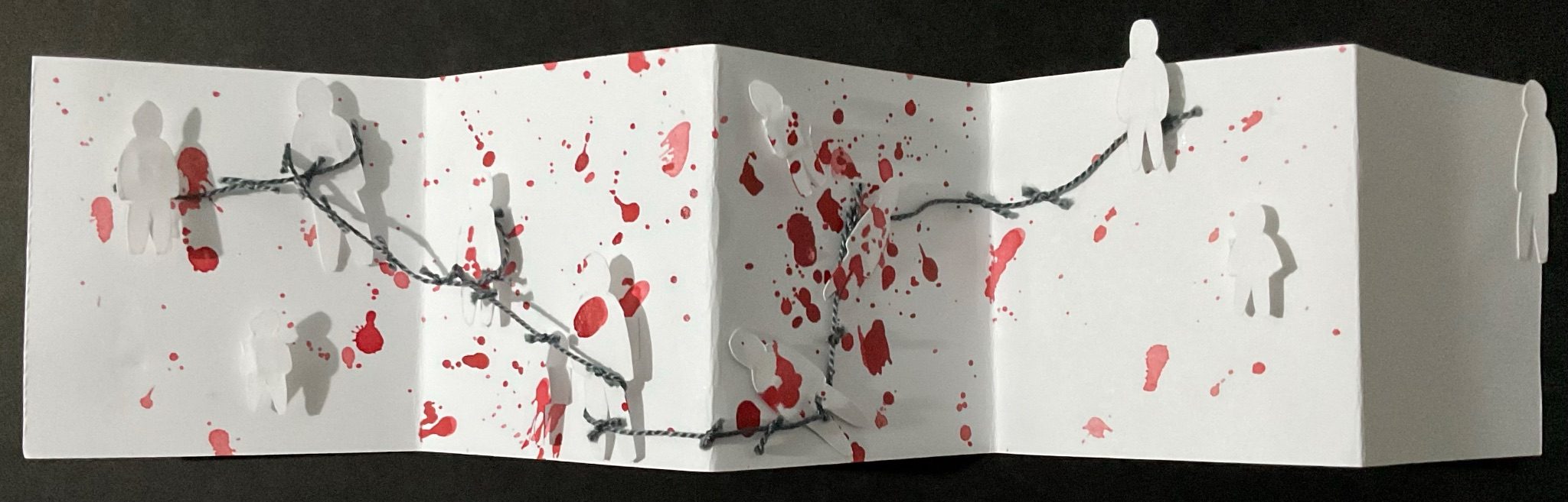 Accordion book. White panels splattered with red ink. Small white figures of different sizes, stand, tilt or collapse. Dark grey thread suggests barbed wire. Front cover, not visible in photo, tiny figure entering. Back cover, slightly larger figure leaving. Evokes fragility, passage and the human cost of violence.