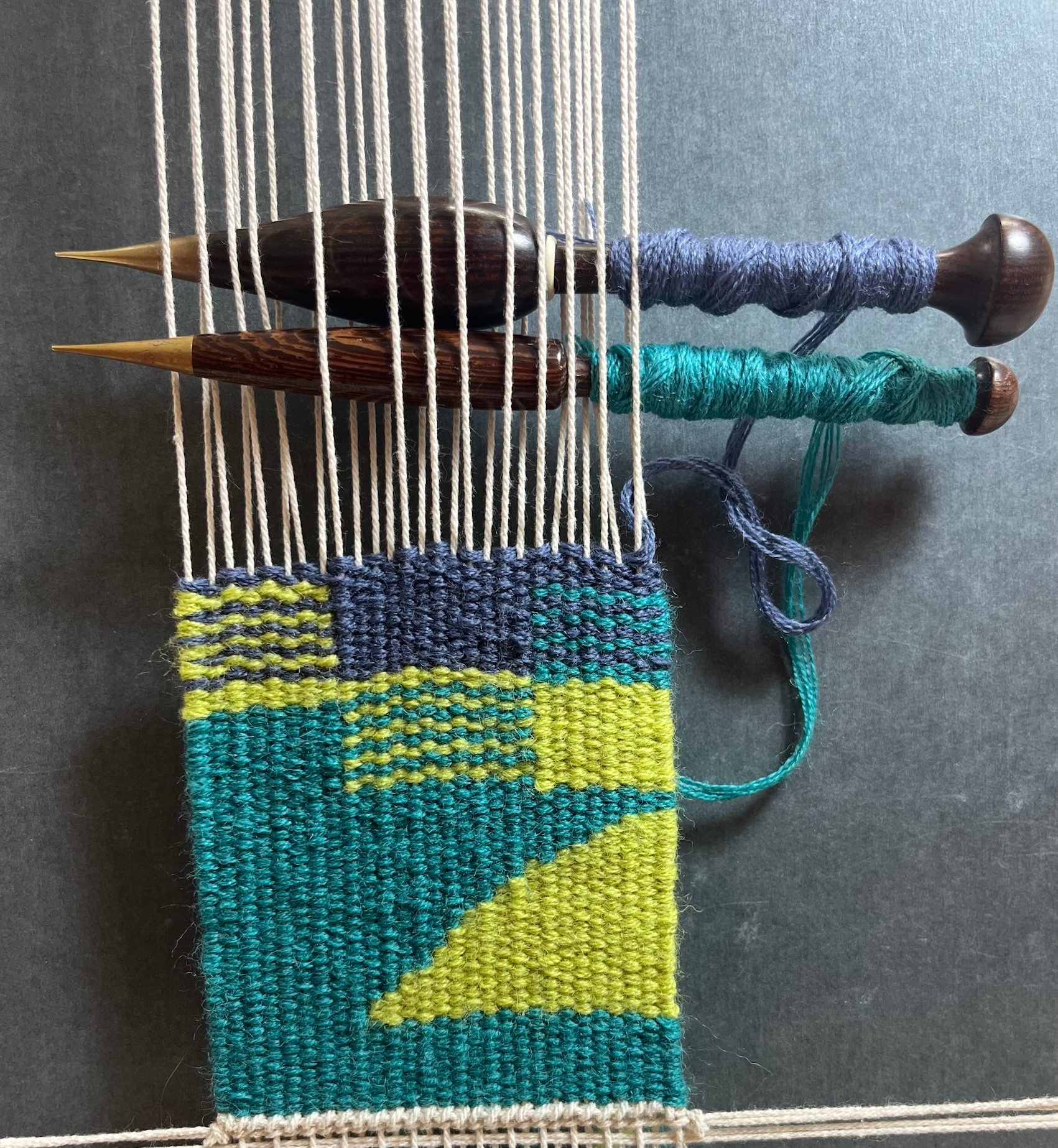 A small tapestry weaving with grey, luminous green and teal blocks of colours as well as stripes.  The tapestry is still on the loom with two wound bobbins at the top.