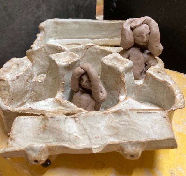 2 clay people curled up in a ceramic egg box.