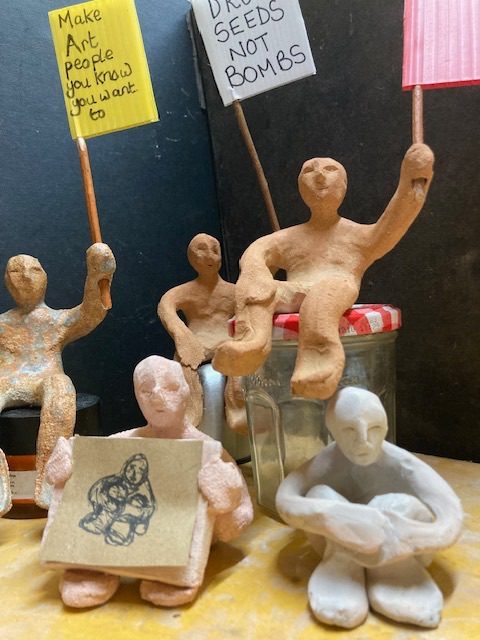5 clay people, 3 with flags, and 2 sitting, one holding a drawing of the other.