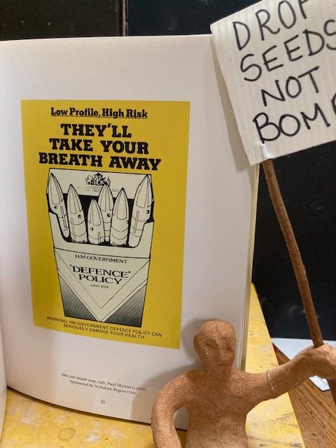 A clay person holding a placard sits in front of a book open at page 37 commenting on the government’s defence policy