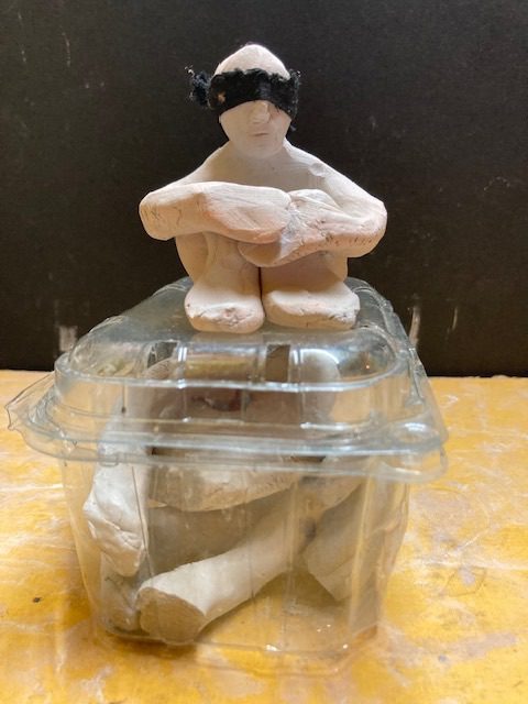 Blindfolded clay person sitting on a plastic container filled with a broken clay person