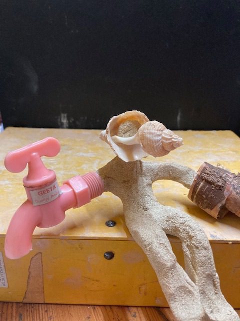 Clay person with a shell as a head, and a pink tap as an arm