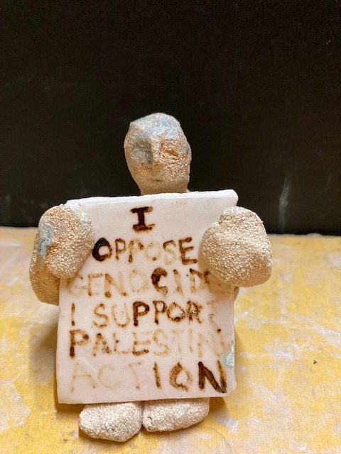 Clay person sitting down holding a placard saying I oppose genocide I support Palestine action