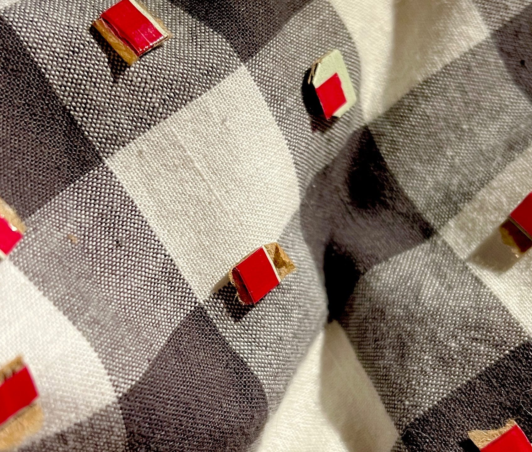 Background of dark grey and white gingham fabric. Tiny squares - red plastic on brown cardboard - lie on the surface. The squares are cut from the ripping strips from card envelopes.