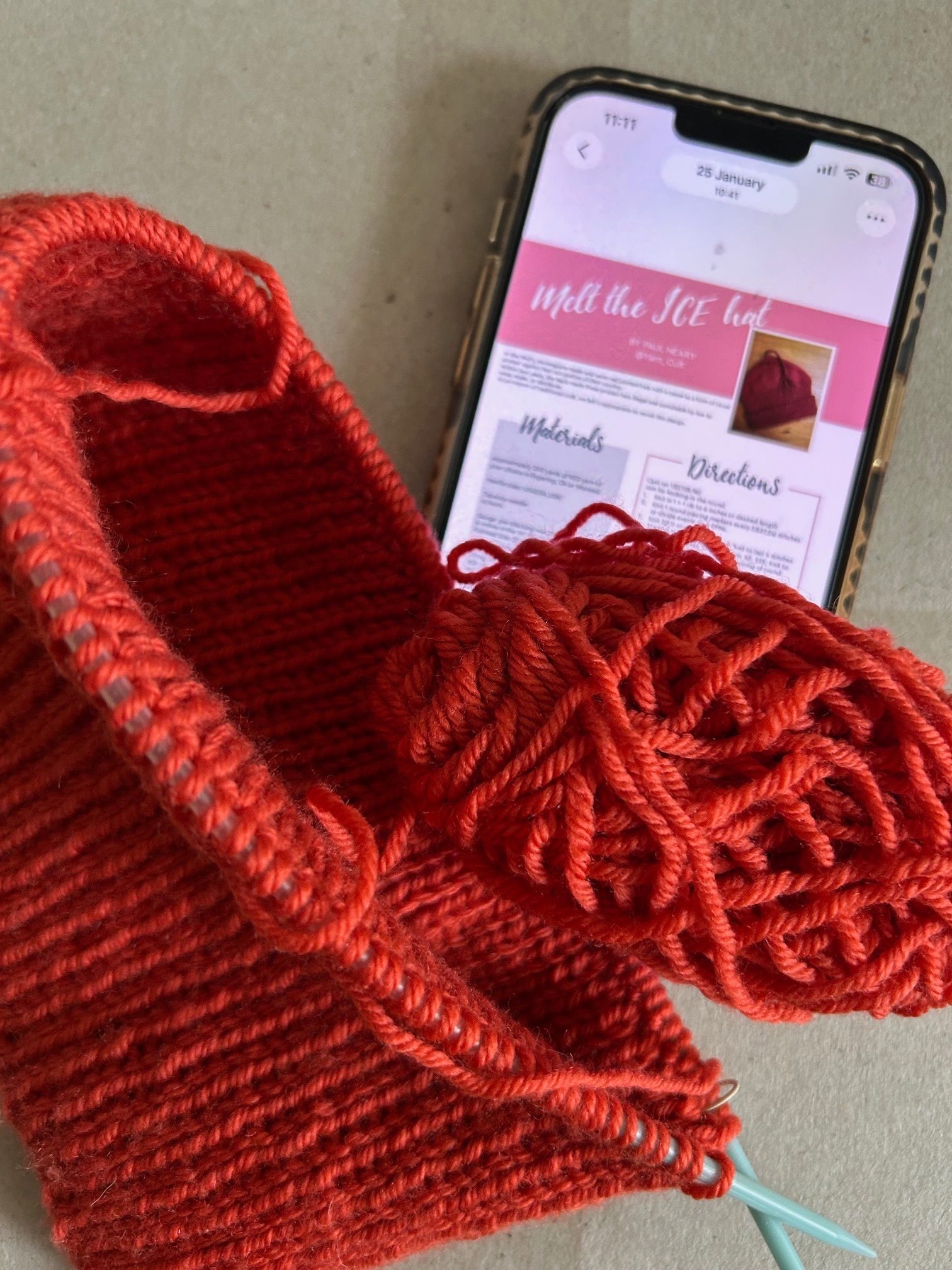 Photograph of knitting with “Melt the ICE” hat pattern on phone in the background.